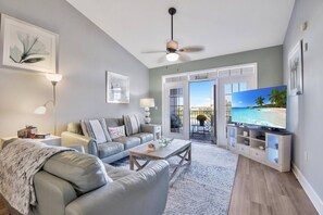 A modern living room with a grey sofa, a coffee table, and a television displaying a beach scene. - Atlantis 302D - Steps to the Ocean (St Augustine Beach)