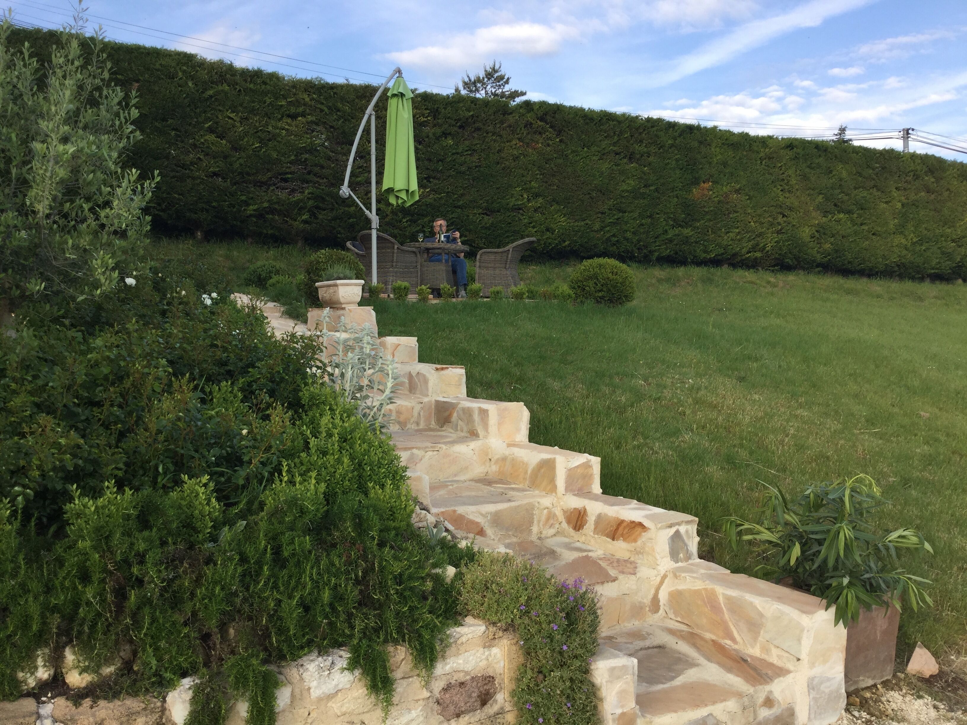 A stone staircase leading up to a patio area with a green umbrella, surrounded by lush greenery and a well-maintained lawn.