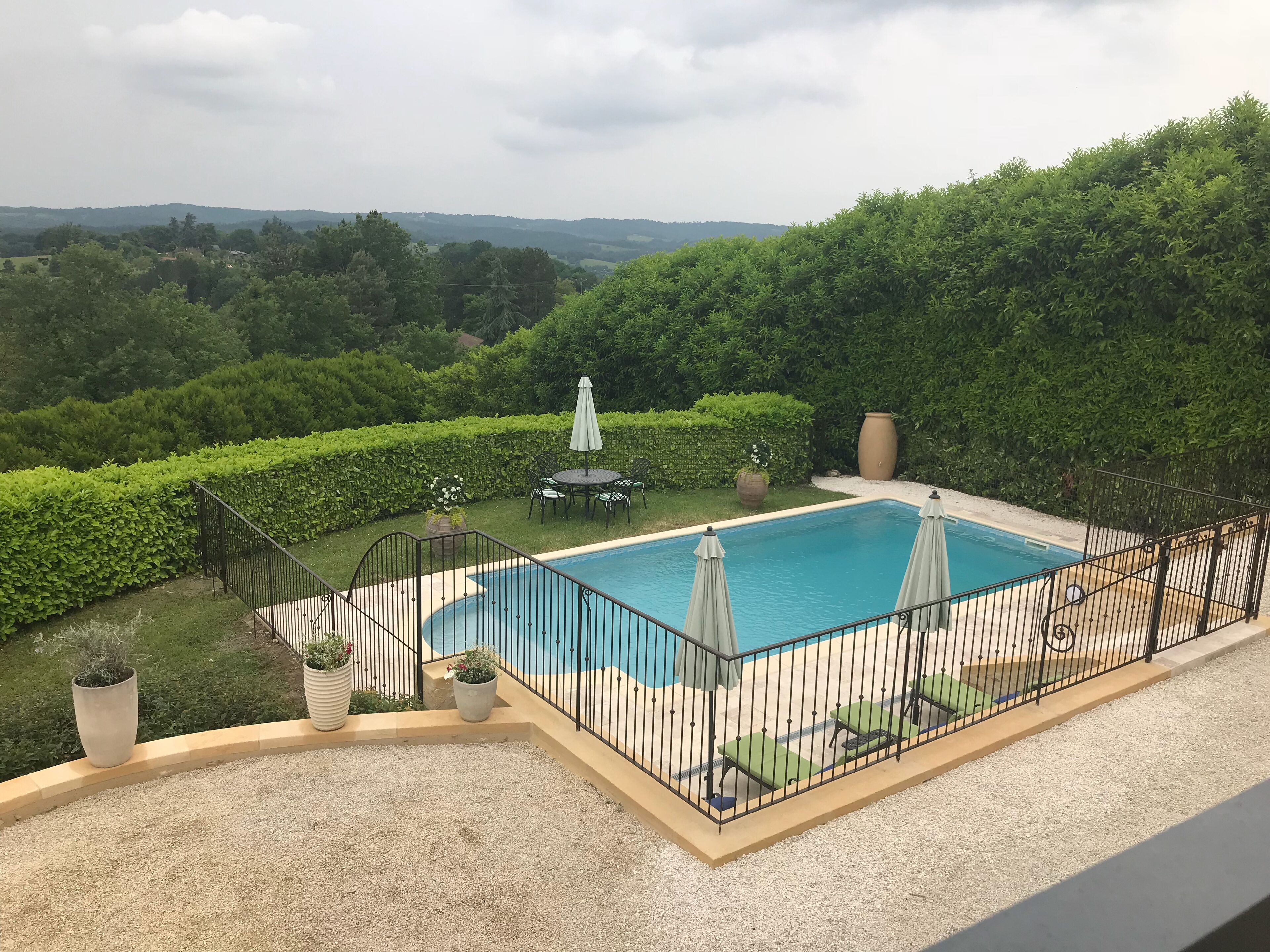 A rectangular pool with a black metal fence, surrounded by a gravel deck. There are two umbrellas and a small table with chairs. The pool is set against a backdrop of lush greenery and distant hills.