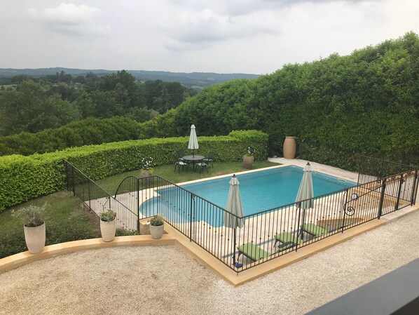 A rectangular pool with a black metal fence, surrounded by a gravel deck. There are two umbrellas and a small table with chairs. The pool is set against a backdrop of lush greenery and distant hills. - Views, air conditioned, heated pool, chefs kitchen,Saturday check-in out (Plazac)