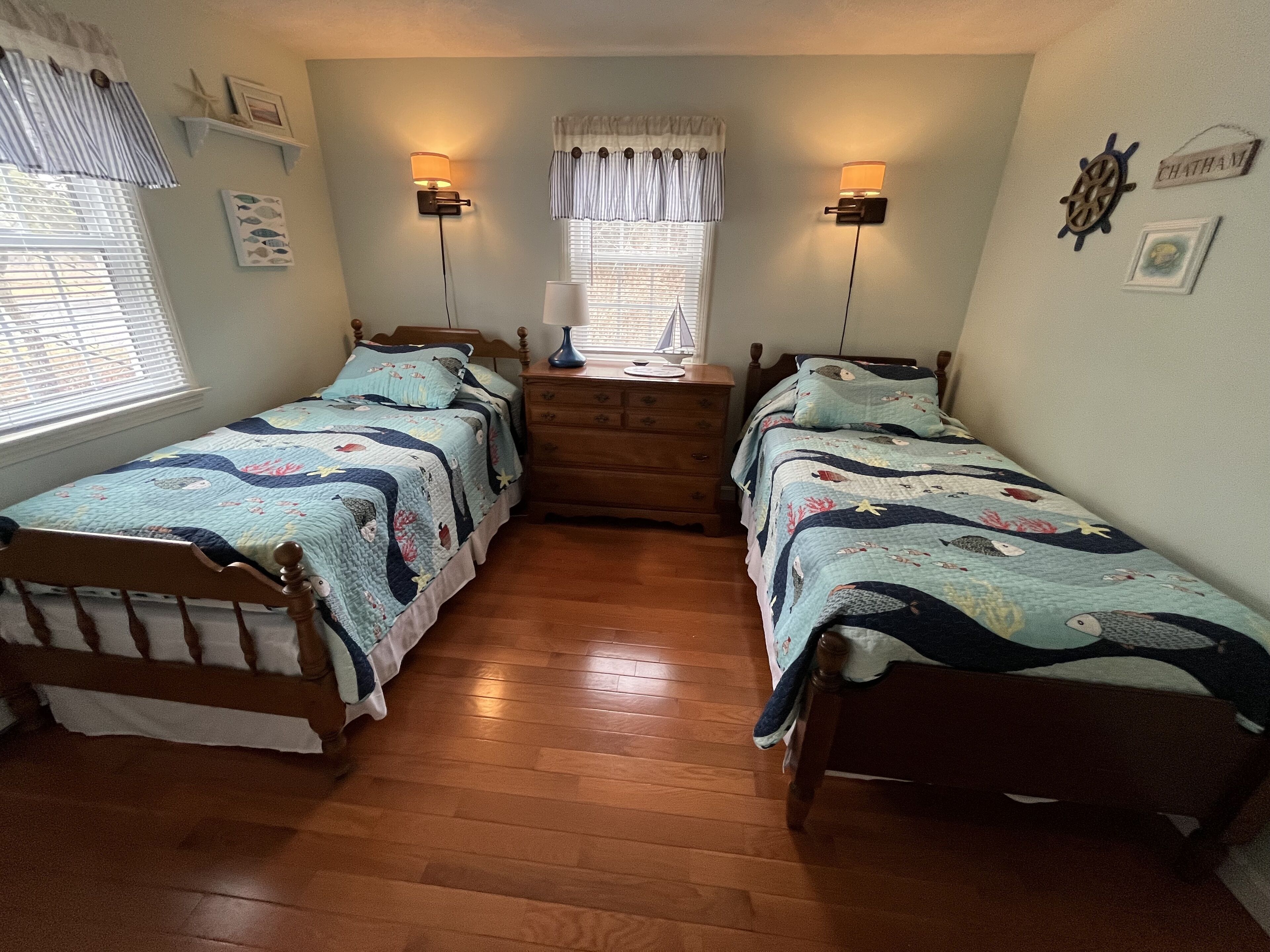A bedroom with two single beds, wooden flooring, a dresser, wall-mounted lamps, and nautical-themed decor.