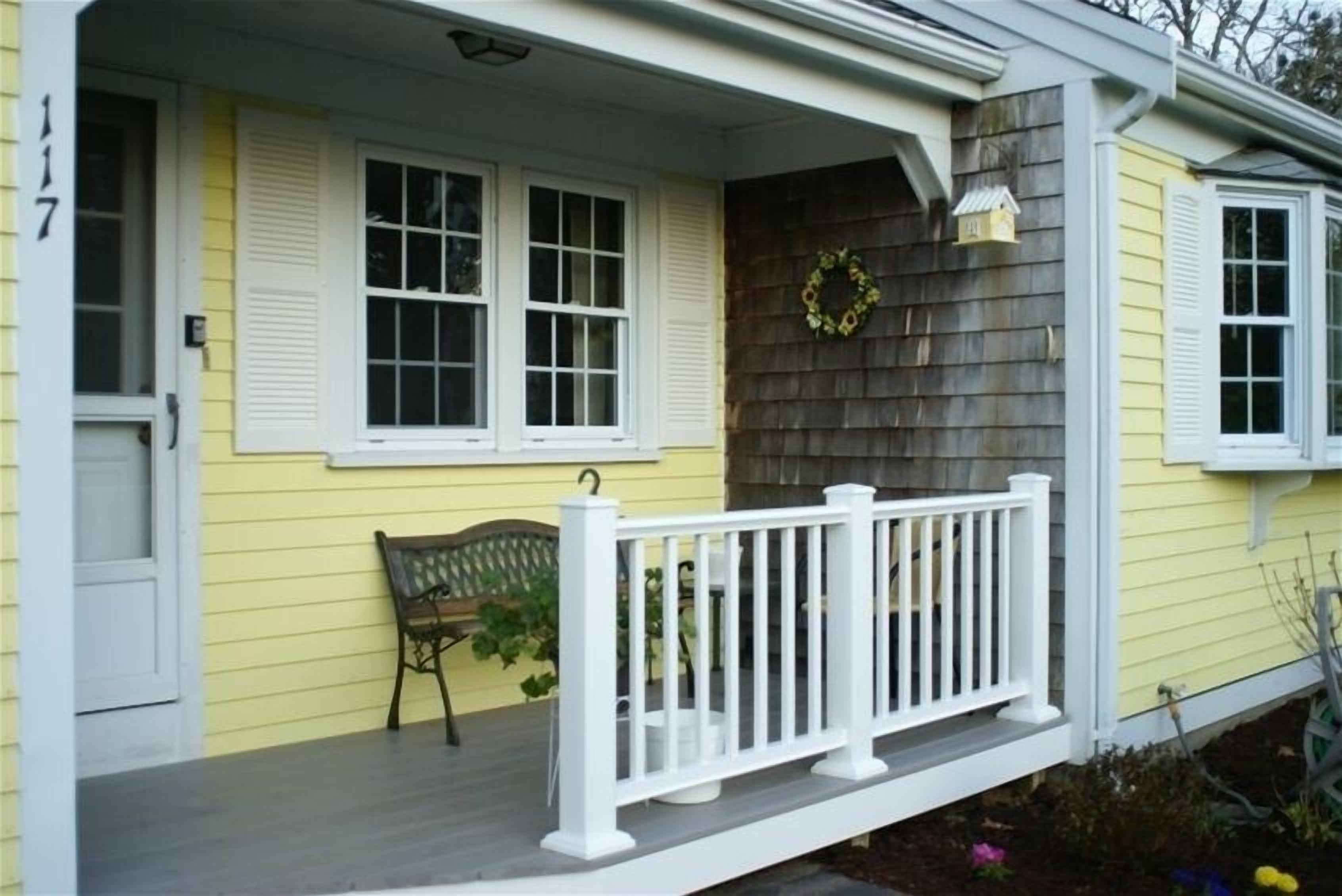 A yellow house with a porch, white shutters, a white railing, and a wreath on the wall.