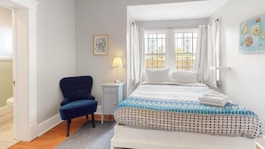 Ein Schlafzimmer mit Bett, Sessel, Nachttisch, Lampe und einem Fenster mit Vorhängen.