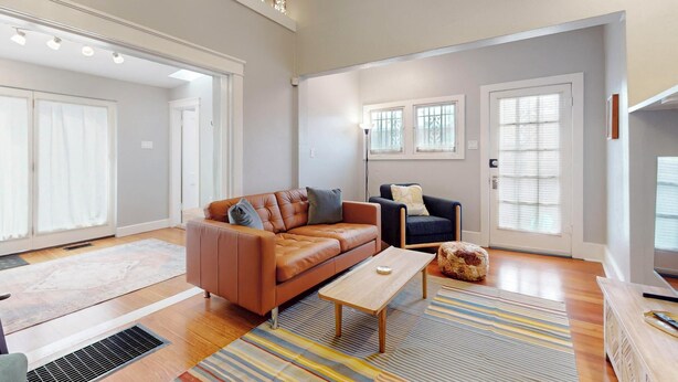 Ein modernes Wohnzimmer mit einem braunen Ledersofa, einem blauen Sessel und einem Holztisch. Große Fenster mit Jalousien, eine Heizung und ein kleiner Teppich sind ebenfalls vorhanden.