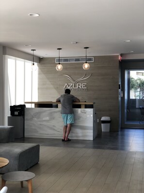 Un comptoir d’accueil au design moderne, le logo d’Azure Beach Residences, et une personne debout derrière. Deux lampes suspendues sont placées au-dessus du comptoir, une poubelle se trouve à droite, et un espace détente avec un canapé et une table ronde est visible au premier plan.