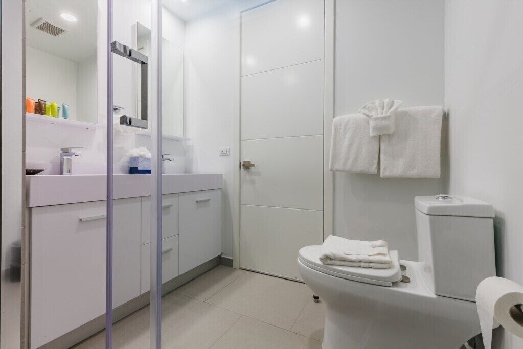 Un bagno moderno con lavandino bianco, specchio e servizi igienici. Asciugamani appesi al muro e cabina doccia in vetro.