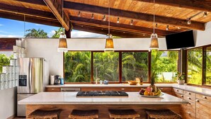 A modern kitchen with a large island, bar stools, a refrigerator, and a view of lush greenery. - Tiki Home - Unique Custom Home Steps to the Beach and Town (Hanalei)