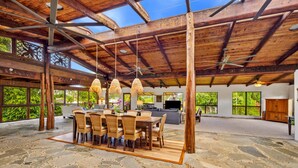 A spacious indoor area with a stone floor, wooden ceiling beams, and large windows offering views of greenery. - Tiki Home - Unique Custom Home Steps to the Beach and Town (Hanalei)