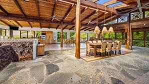 A spacious interior with a stone floor, wooden ceiling beams, and large windows offering a view of greenery. - Tiki Home - Unique Custom Home Steps to the Beach and Town (Hanalei)
