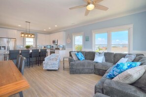 A modern kitchen with white cabinets, a dining area with a wooden table, a gray sectional sofa with blue pillows, and a ceiling fan.