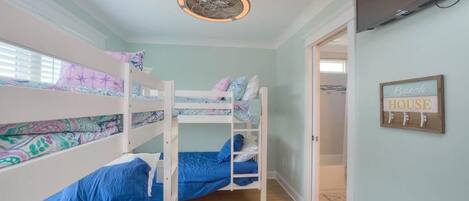 A bunk bed room with a ceiling fan, a door leading to another room, and a sign on the wall that says "Beach HOUSE".