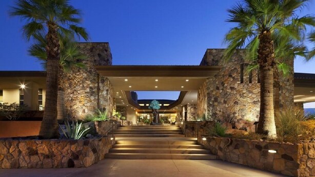 A hotel entrance with a stone facade, palm trees, and a covered walkway leading to the main building.