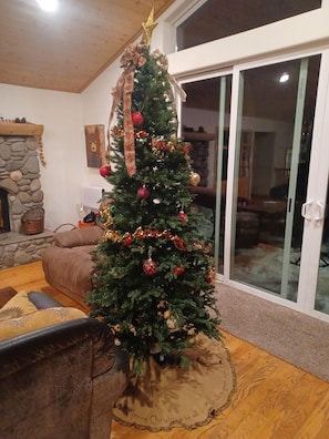 Un salón acogedor con un árbol de Navidad decorado, una chimenea de piedra y un gran ventanal corredizo que da a un balcón.