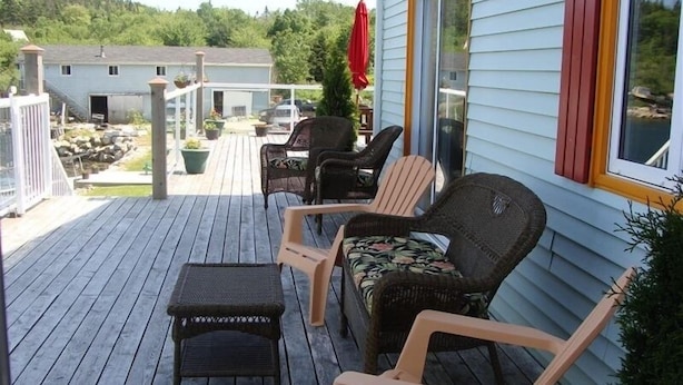 Una terraza de madera con mobiliario exterior, incluyendo sillas y una pequeña mesa, y una casa con un paraguas rojo.