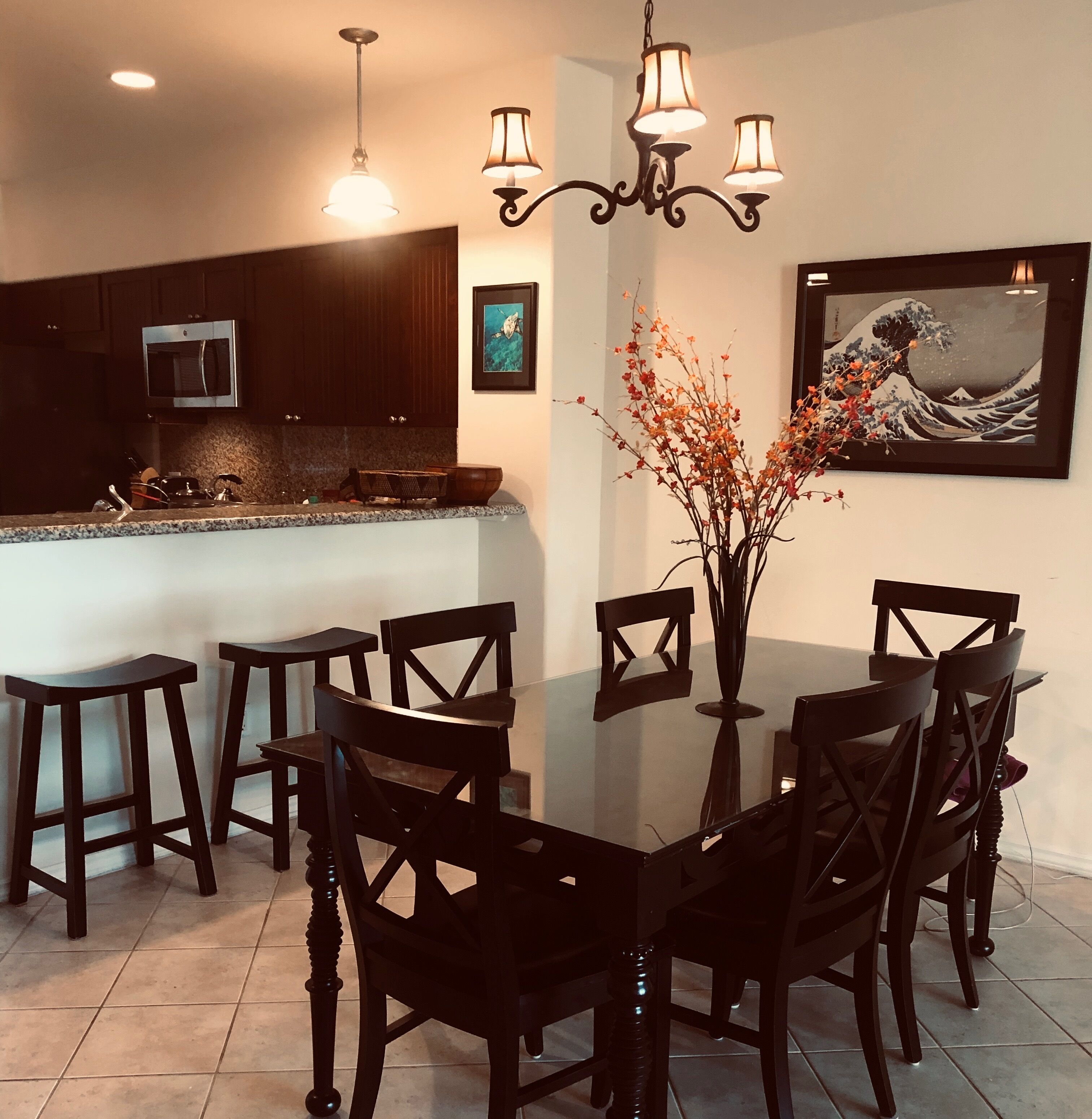 A dining area with a glass table surrounded by chairs, a chandelier, a kitchen in the background, and a framed artwork on the wall.