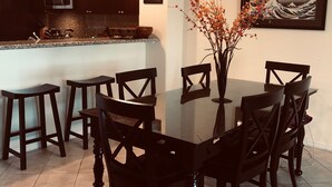 Un comedor con una mesa de vidrio rodeada de sillas, una araña, una cocina al fondo y una obra de arte enmarcada en la pared.