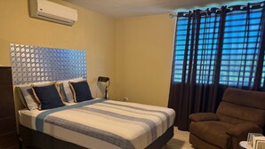 Ein Schlafzimmer mit einem Bett, einem braunen Sessel, einem kleinen Tisch und einem Fenster mit Vorhängen.