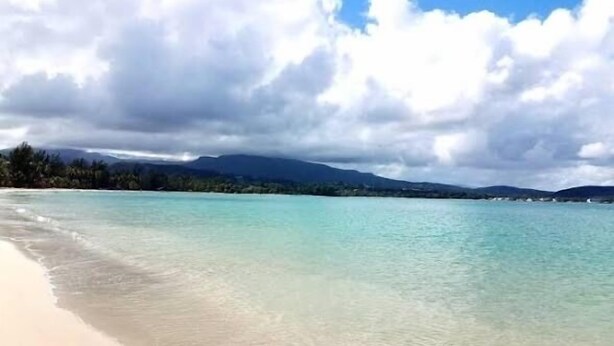 Ein Strand mit klarem türkisblauem Wasser, einem Sandstrand und einem Hintergrund aus Bergen und Wolken.