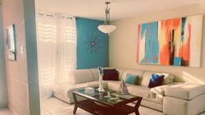 Ein Wohnzimmer mit einem weißen Sofa, einem Glastisch und einem farbenfrohen, abstrakten Gemälde an der Wand.