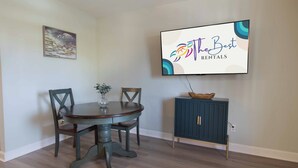 A dining area with a round table, four chairs, a vase with flowers, a cabinet, and a television displaying 'The Best Rentals' logo. - 1Br/1Ba Close to Beach & Shops- Heron's Nest (Folly Beach)