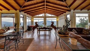 A spacious living area with a high wooden ceiling, a dining table with chairs, a sofa, and a coffee table. There are large windows offering a view of the mountains. - Heated Infinity Pool Villa, 6BR, 25Min SJO (Alajuela)