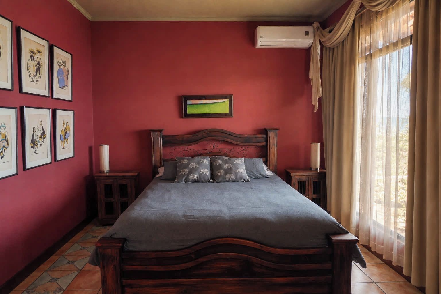 A bedroom with a bed, a wall-mounted air conditioner, a window with curtains, and framed pictures on the wall.