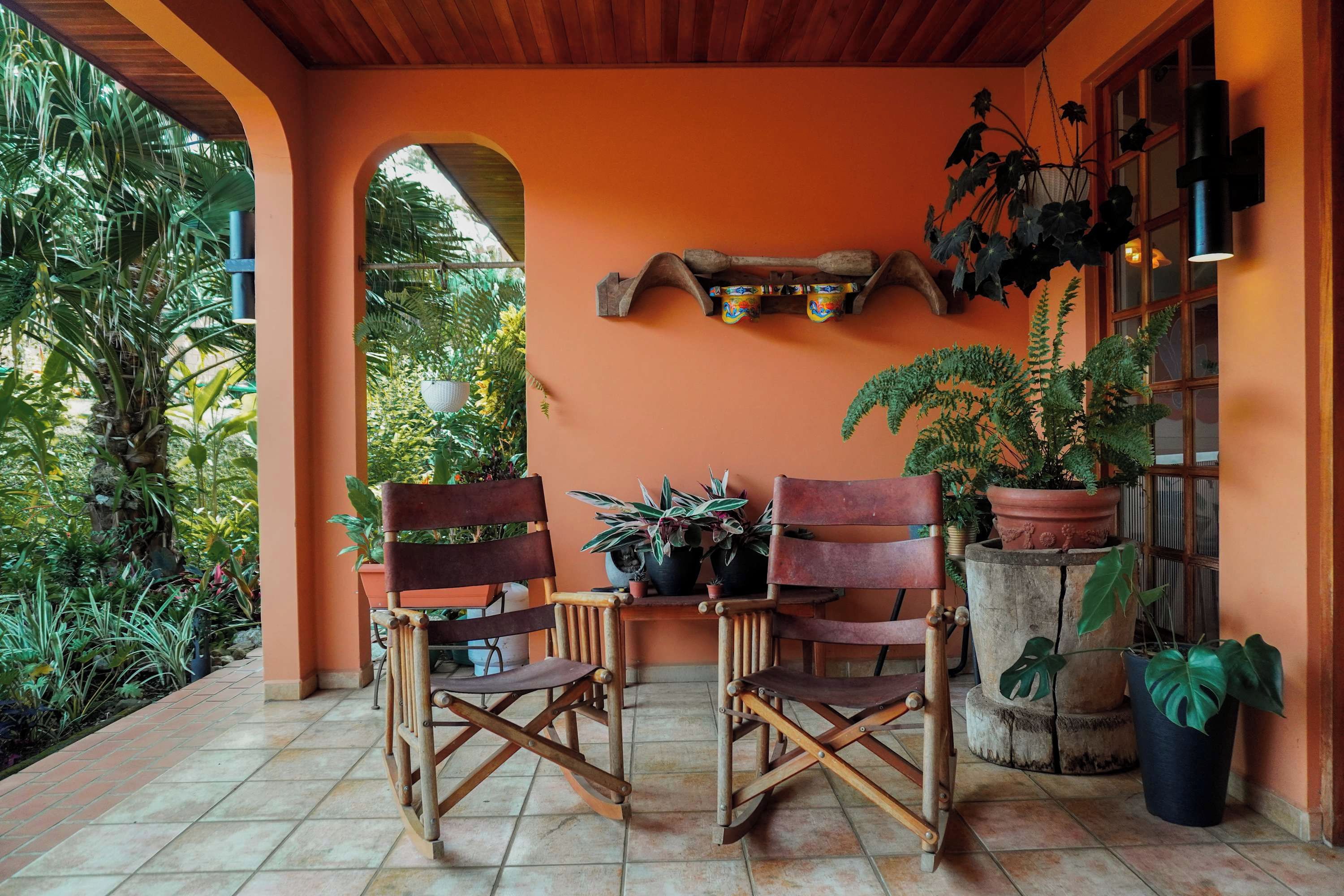 A covered outdoor seating area with two wooden chairs, a small table, and various potted plants.