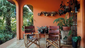 A covered outdoor seating area with two wooden chairs, a small table, and various potted plants.