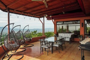 An outdoor patio with a table and chairs, a hanging chair, and a view of the city and mountains. - Heated Infinity Pool Villa, 6BR, 25Min SJO (Alajuela)