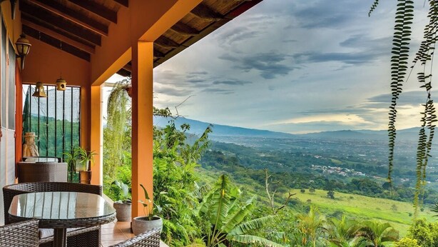 A covered outdoor seating area with wicker furniture, potted plants, and a view of a lush valley. - Heated Infinity Pool Villa, 6BR, 25Min SJO (Alajuela)