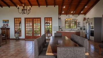 A spacious dining area with a long wooden table and grey upholstered chairs, a kitchen with modern appliances, and a wooden ceiling.