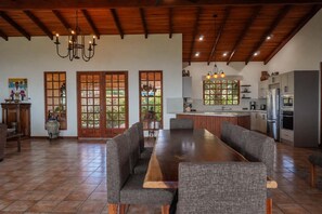 A spacious dining area with a long wooden table and grey upholstered chairs, a kitchen with modern appliances, and a wooden ceiling. - Heated Infinity Pool Villa, 6BR, 25Min SJO (Alajuela)