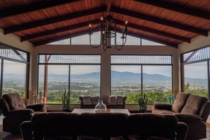 A spacious room with a high ceiling, large windows offering a view of a cityscape and mountains, and a chandelier. - Heated Infinity Pool Villa, 6BR, 25Min SJO (Alajuela)