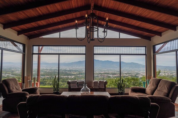 Una habitación amplia con techo alto, ventanas grandes que ofrecen vistas a un paisaje urbano y montañas, y un candelabro.