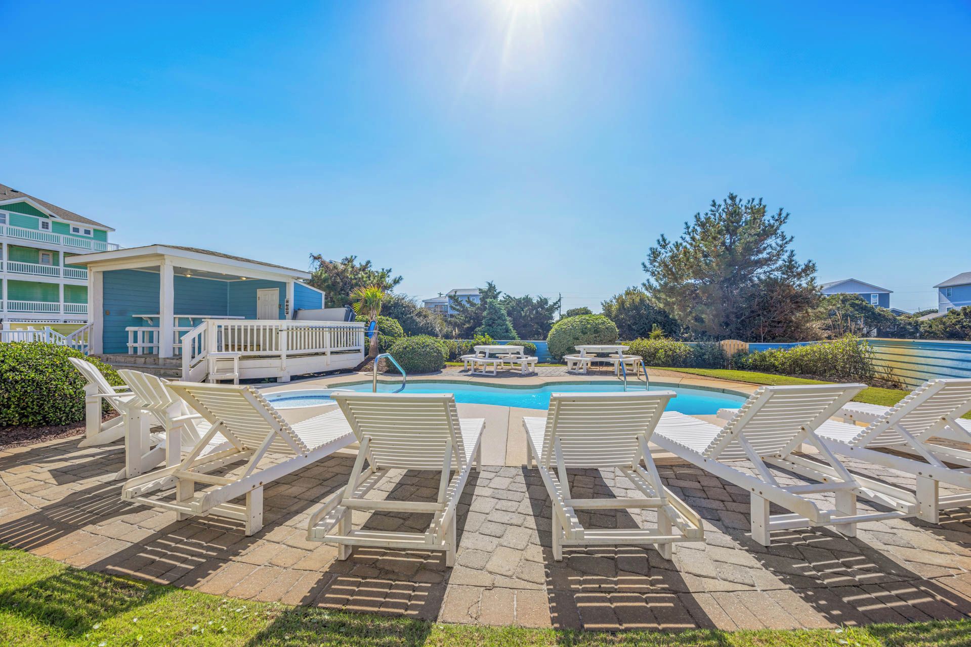 The property features a sunny poolside area with white lounge chairs arranged on a paver deck, surrounded by lush green lawns, mature trees, and a peaceful coastal neighborhood setting.