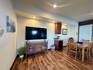 A living room with a flat-screen TV, wooden cabinet, dining table with chairs, and a potted plant.