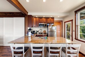 Una cocina con encimera de granito, sillas blancas, armarios de madera, electrodomésticos de acero inoxidable y una ventana con vistas a la vegetación.