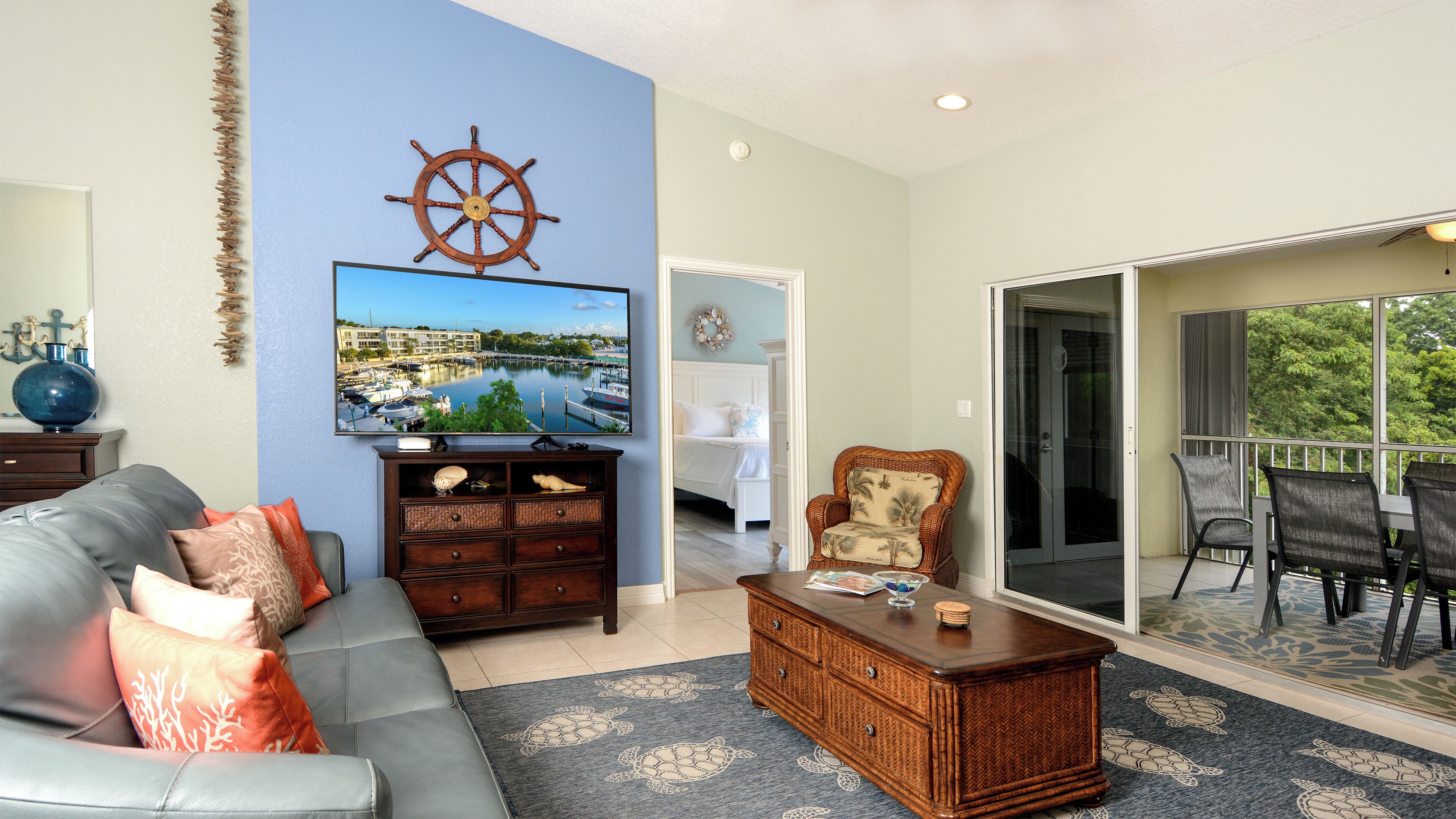 A living room with a sofa, a coffee table, a television, a wall-mounted ship's wheel, and a view through a glass door to an outdoor patio.