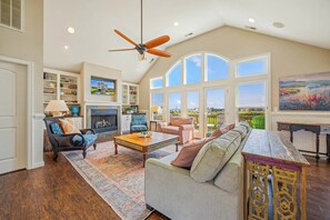 A spacious living room with a fireplace, large windows, and a ceiling fan. - Oceanview Home w/ Private Pool, Hot Tub, and Elevator. Enjoy the HI View! (Rodanthe)
