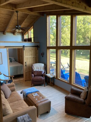 A cozy room with a bed, a brown leather chair, a brown leather sofa, and a small table. There is a large window with a view of outdoor furniture.