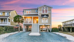 A two-story beachfront house with a balcony, a covered porch, and a staircase leading to the main entrance. - Luxury Oceanfront Retreat: Concierge Beach Tent Service, Private Pool & Elevator (Holden Beach)