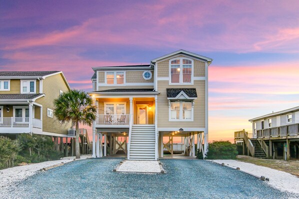 A two-story beachfront house with a balcony, a covered porch, and a staircase leading to the main entrance. - Luxury Oceanfront Retreat: Concierge Beach Tent Service, Private Pool & Elevator (Holden Beach)