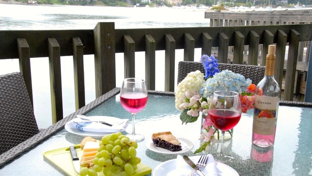 A table set with a bottle of wine, glasses, a plate of cheese, grapes, a pie, and a vase of flowers. -  Unique Marina Side Condo With Great Views And Fun Atmosphere! (Southwest Harbor)