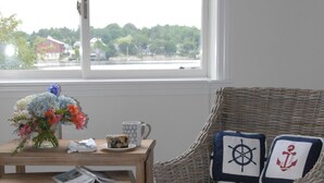 A wicker chair with nautical-themed cushions, a small wooden table with a vase of flowers, a mug, and a red ottoman in a room with a window overlooking a green landscape. -  Unique Marina Side Condo With Great Views And Fun Atmosphere! (Southwest Harbor)