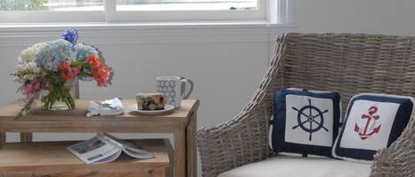 Une chaise en osier agrémentée de coussins aux motifs marins, une petite table en bois sur laquelle est posé un vase de fleurs, une tasse et un pouf rouge, le tout dans une pièce dotée d’une fenêtre donnant sur un paysage verdoyant.