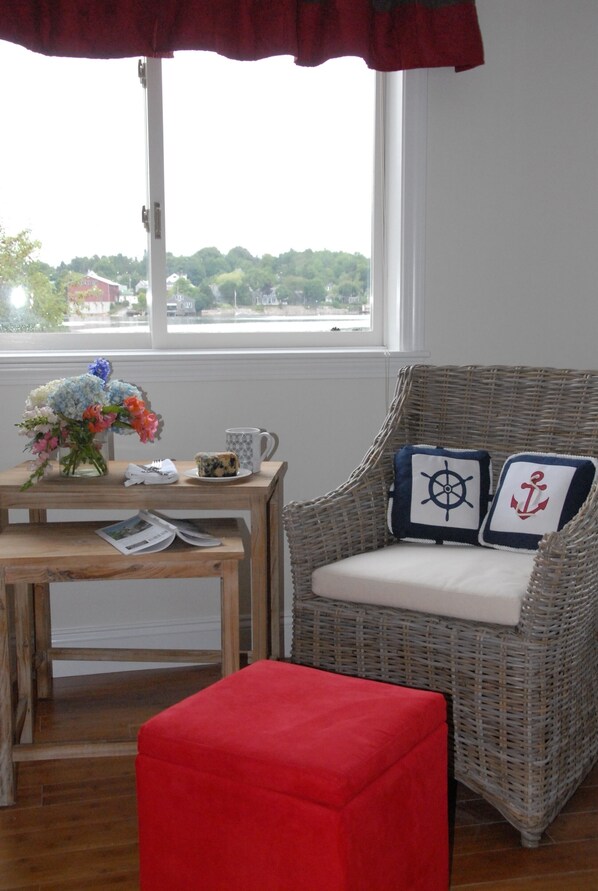 A wicker chair with nautical-themed cushions, a small wooden table with a vase of flowers, a mug, and a red ottoman in a room with a window overlooking a green landscape.