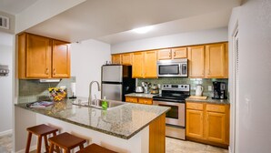 A kitchen with wooden cabinets, a stainless steel refrigerator, a microwave, an oven, and a coffee maker. There are two wooden stools at the island. - SLEEPS 6 Beautiful, Fully Furnished Condo - Great Price, Perfect Location! (Tempe)