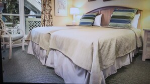 A double bed with a beige bedspread and pillows, a nightstand with a lamp, a chair, and a balcony with a view of a building and trees.