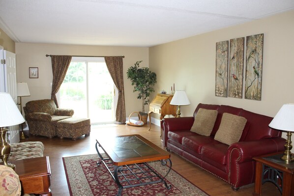 A living room with a red leather sofa, a patterned armchair, a wooden coffee table, and a red rug. There are two lamps, a plant, and artwork on the walls.