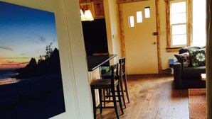 Ein Essbereich mit Holzboden, einem Tisch und Stühlen. Im Hintergrund ist eine Küche mit Holzausbau und einem Fenster zu sehen.